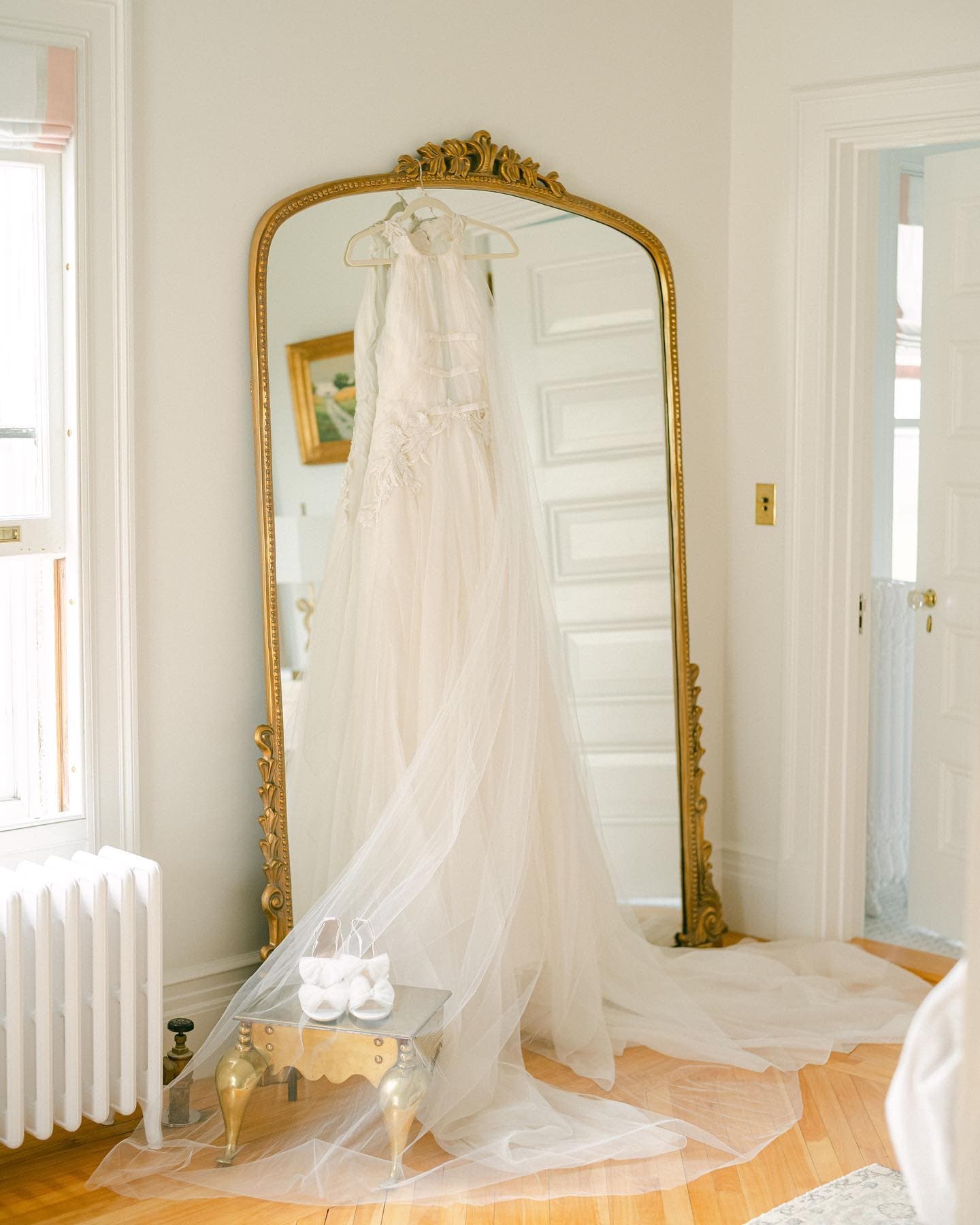 A delicate white wedding dress hangs in front of a gold-framed mirror with soft lighting.