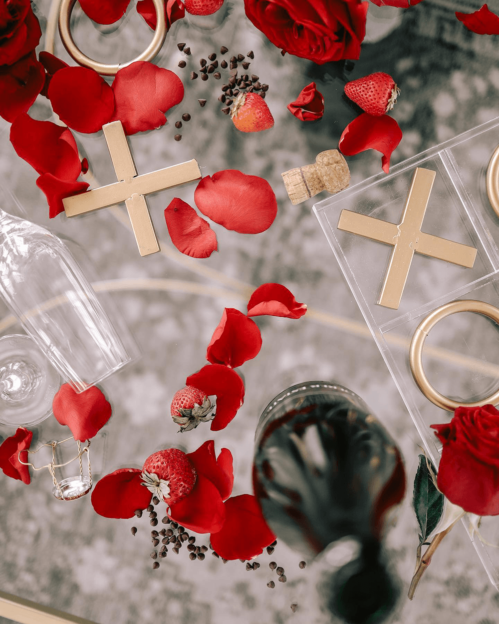 An overhead view of a romantic setup featuring red rose petals, strawberries, gold decorations, and a bottle.