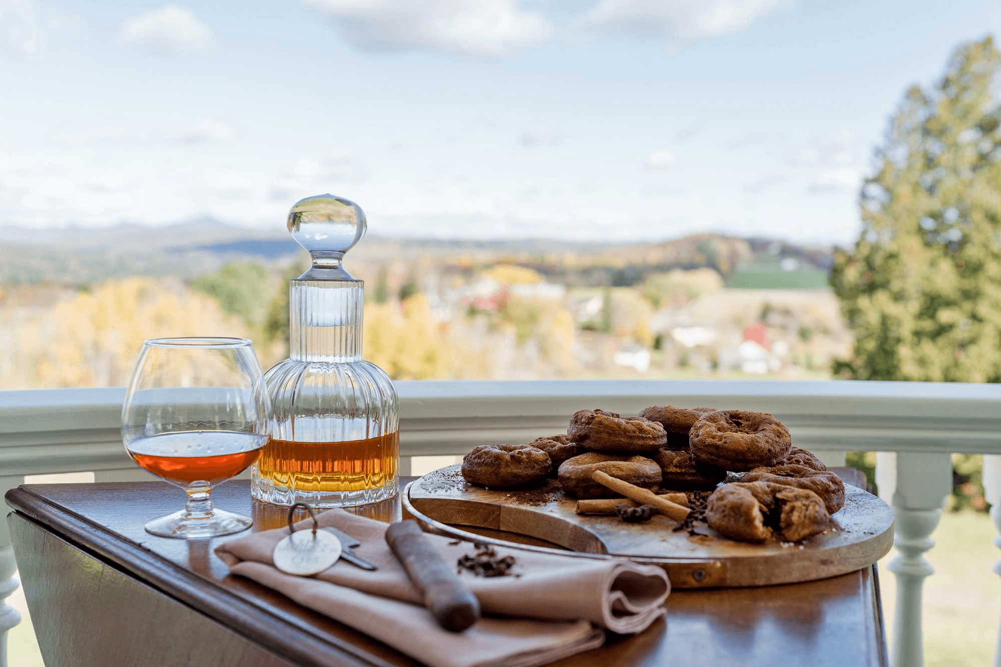 A glass of amber liquor, a decanter, and fresh donuts are arranged on a wooden table overlooking a scenic landscape.