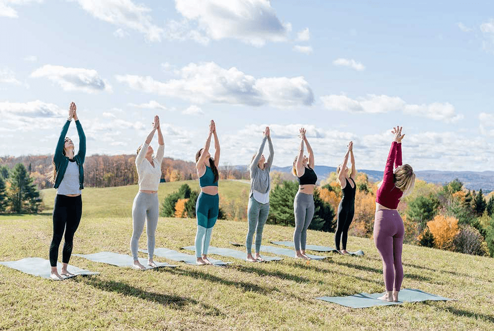 A group of women practicing yoga outdoors, standing on mats with arms raised towards the sky in a scenic landscape.