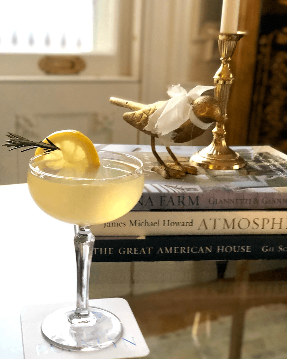 A cocktail garnished with a lemon slice and rosemary sits on a stack of books beside a decorative bird figurine and a candle holder.