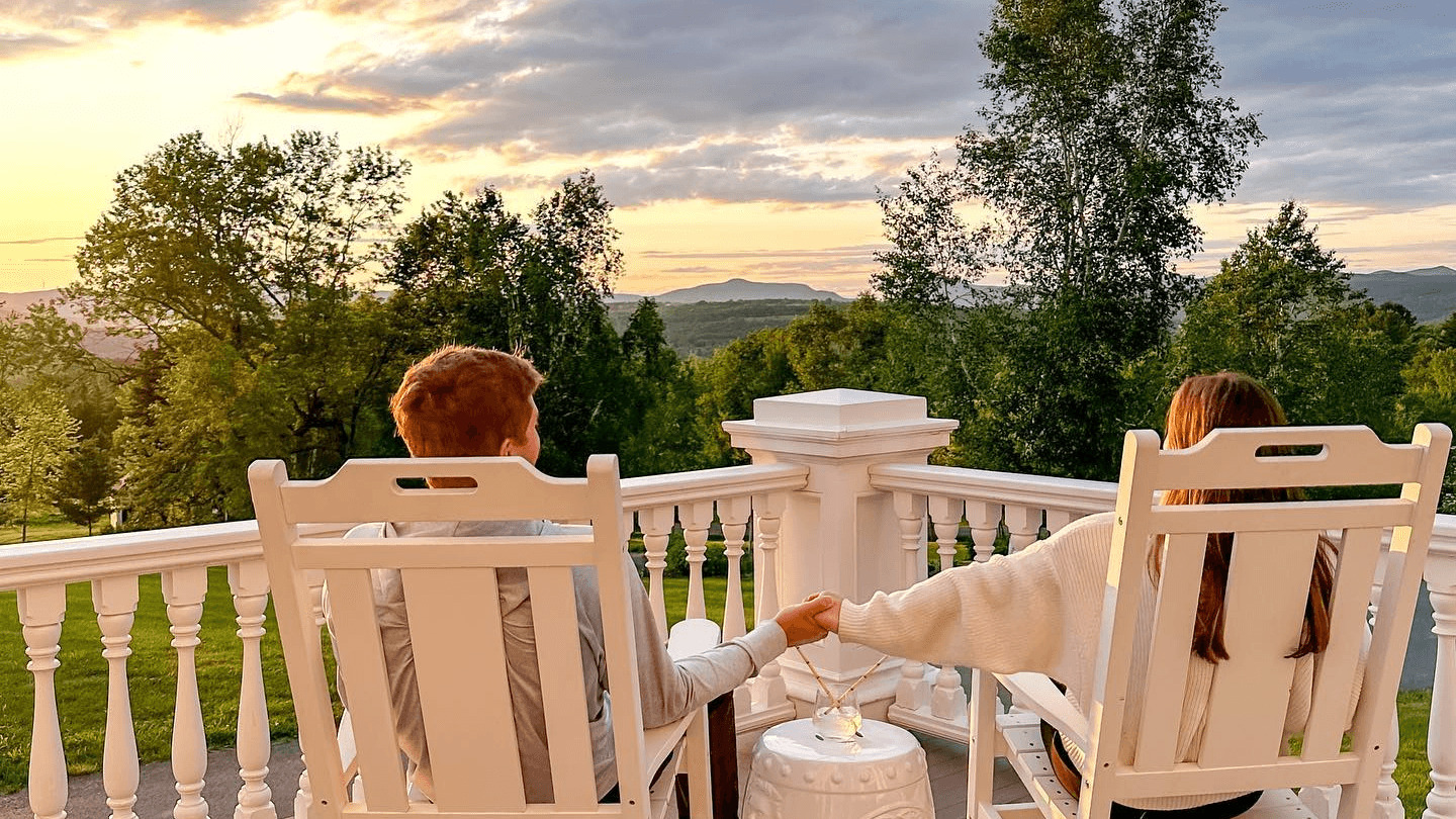 A couple sits hand-in-hand on a porch, enjoying a sunset view of the mountains and trees.