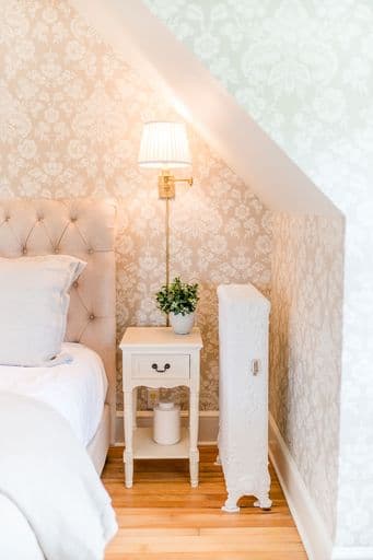 A cozy bedroom corner featuring a tufted headboard, a bedside table with a lamp and a small plant, and a white radiator against patterned wallpaper.
