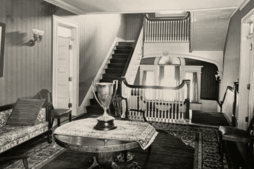 A vintage black-and-white photo of a cozy foyer with a staircase, a patterned rug, a decorative table, and a couch.