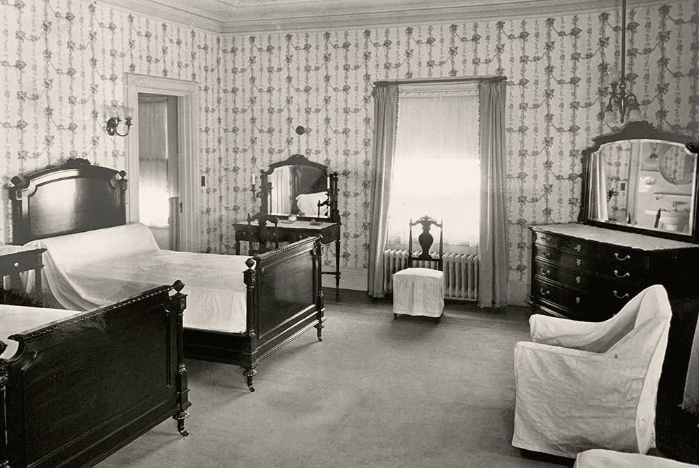 A vintage black-and-white photo of a furnished bedroom featuring two beds, a dresser, and several mirrors against a floral wallpaper backdrop.