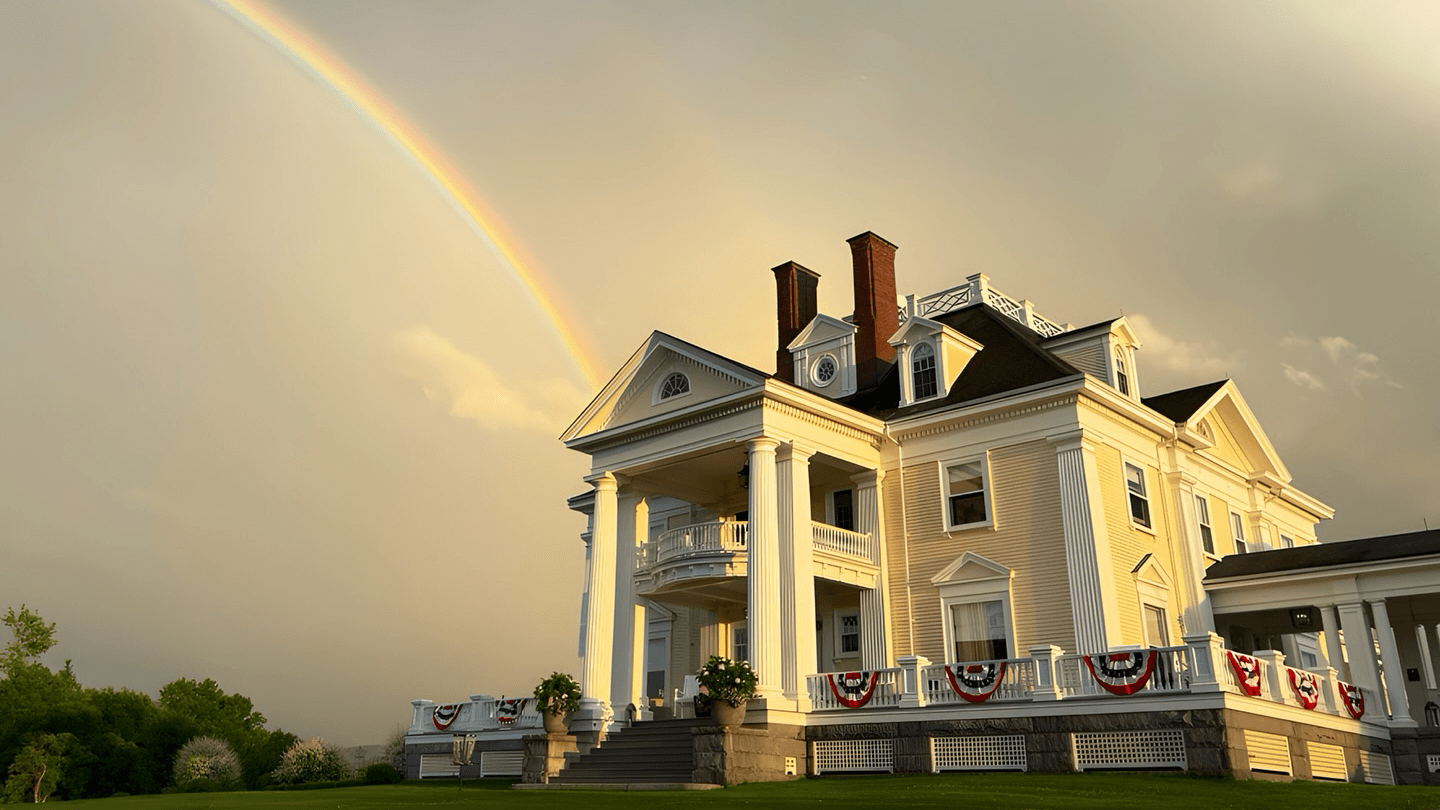 A grand white mansion is set against a backdrop of a rainbow and cloudy sky.
