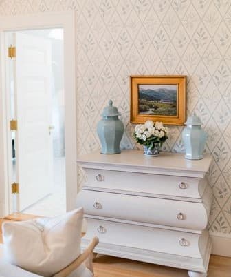 A stylish room featuring a light-colored dresser adorned with two blue vases and a floral arrangement against a patterned wall.
