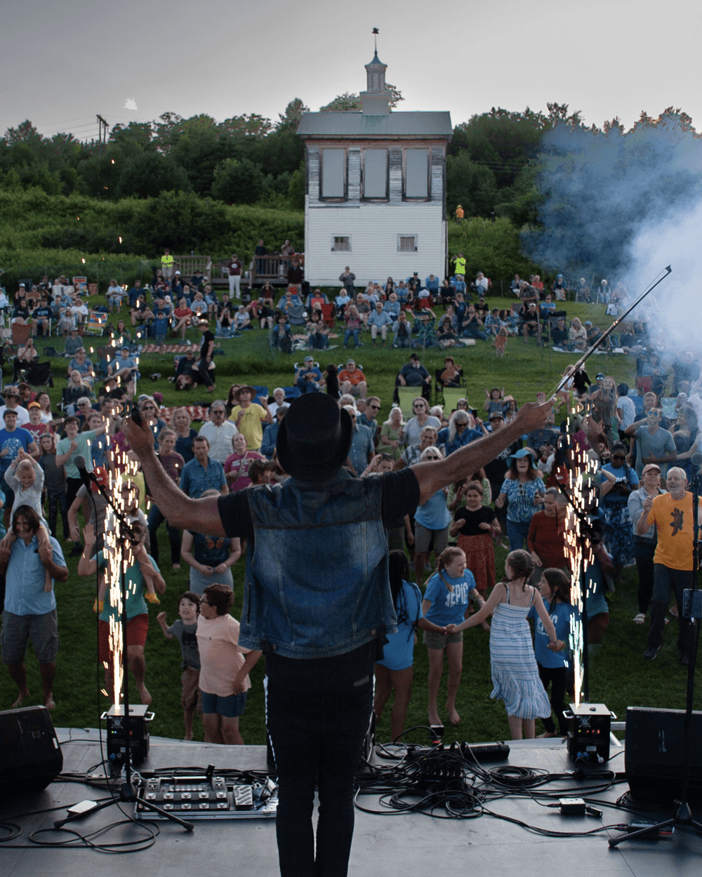 A performer on stage ignites sparks while a large crowd enjoys the outdoor concert in the background.
