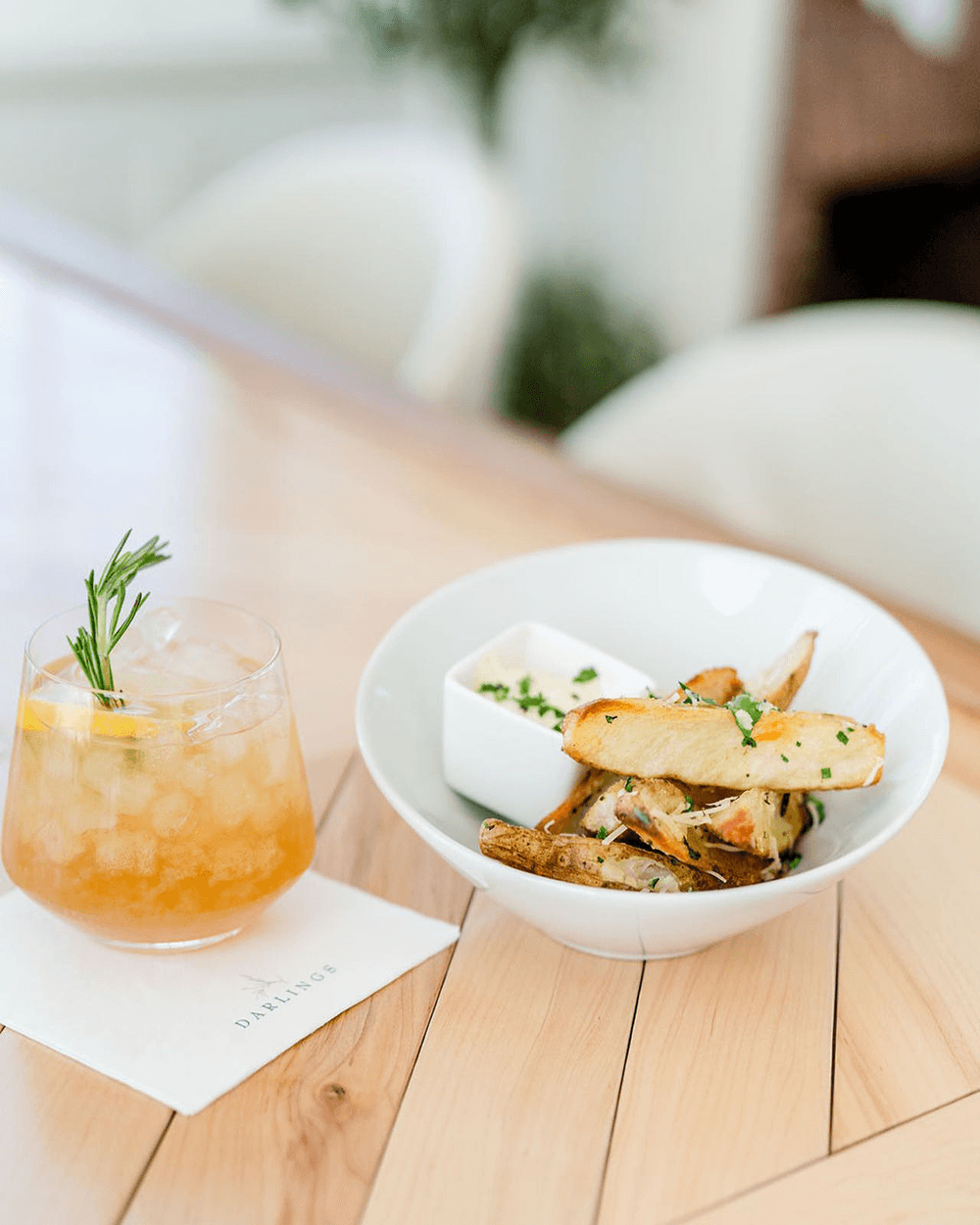 A cocktail and a bowl of crispy potato wedges with dipping sauce on a wooden table.