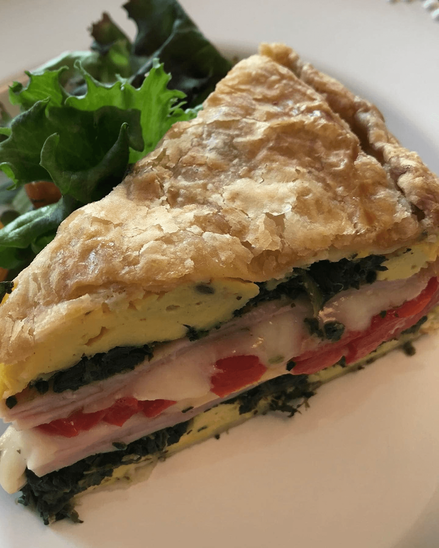 A slice of layered savory pastry filled with greens, ham, cheese, and tomatoes, served with a side of salad.