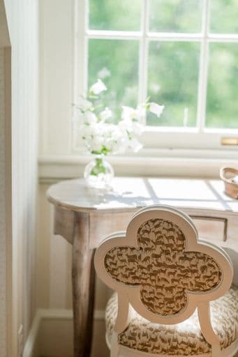 A light-filled room features a wooden table adorned with a vase of white flowers and a decorative chair with a patterned backrest.