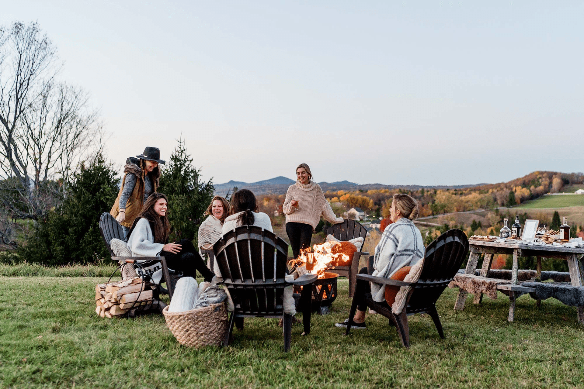 A group of women sits around a fire pit in a scenic outdoor setting, enjoying each other's company.