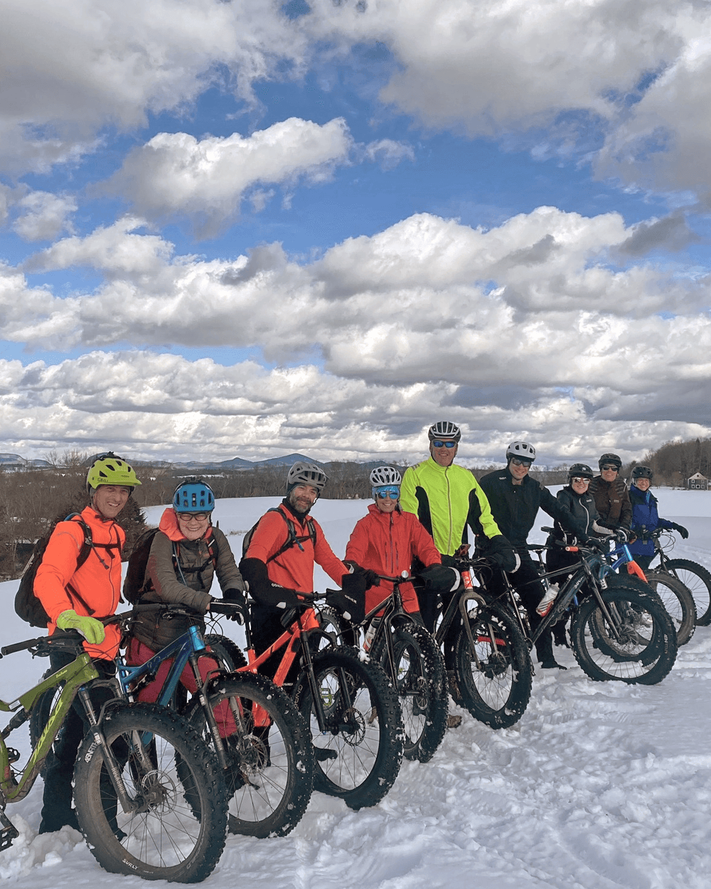 A group of eight people in colorful cycling gear poses on fat bikes in the snow under a partly cloudy sky.