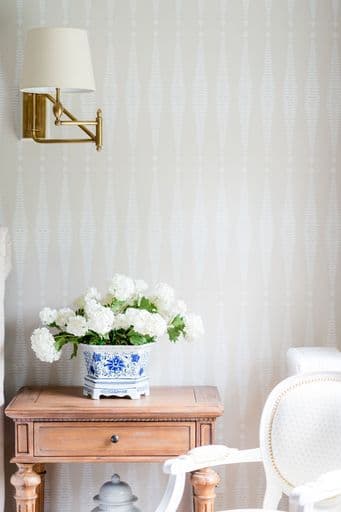 A porcelain vase with white flowers sits on a wooden table next to a gold wall sconce in a softly patterned room.