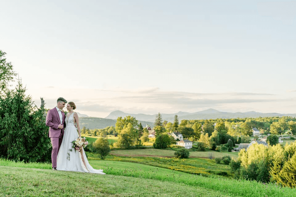 A couple in wedding attire stands together on a grassy hillside with a scenic landscape in the background.