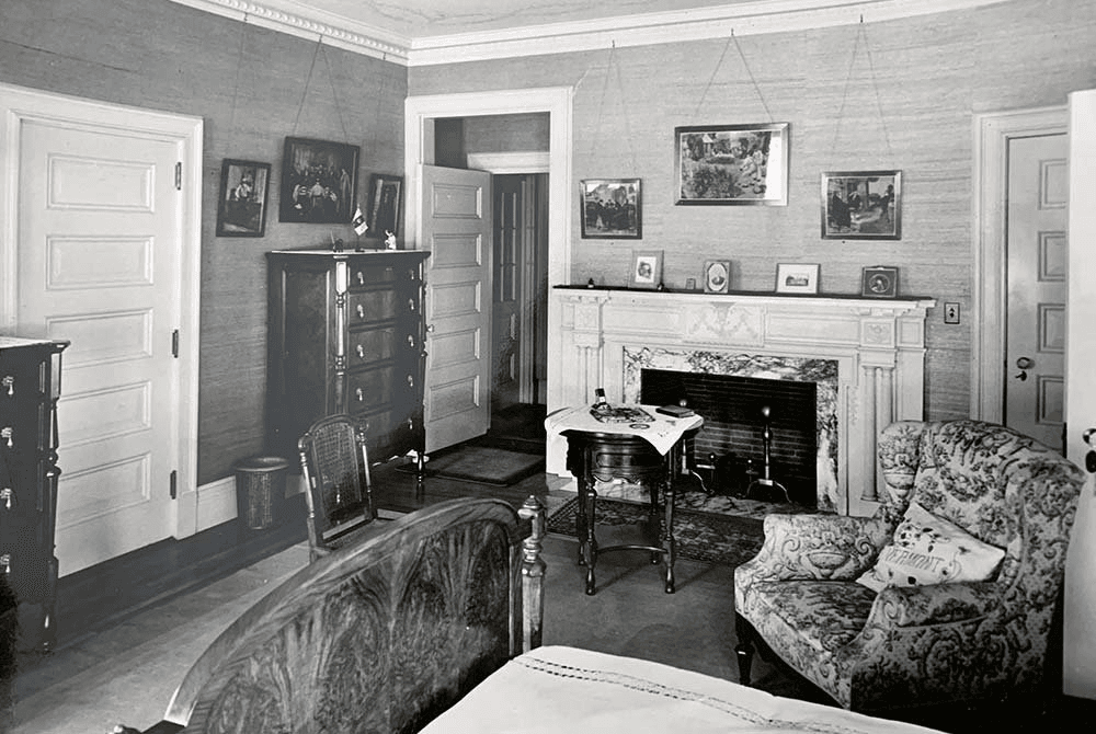 A vintage black-and-white interior photograph of a bedroom featuring a decorative fireplace, patterned armchair, and wooden furniture.