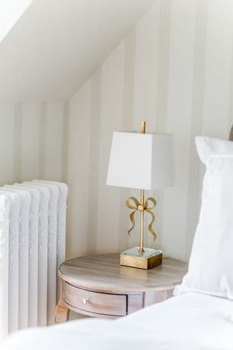 A bedside table featuring a small lamp with a white shade and a decorative gold bow, beside a white radiator.