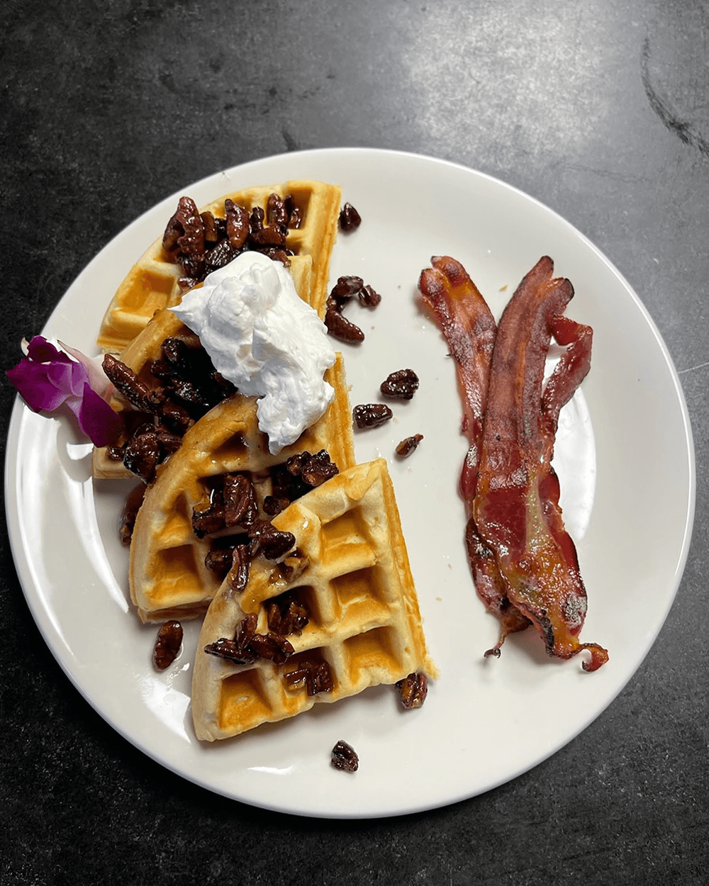 A plate of waffles topped with nuts, whipped cream, and a side of crispy bacon.