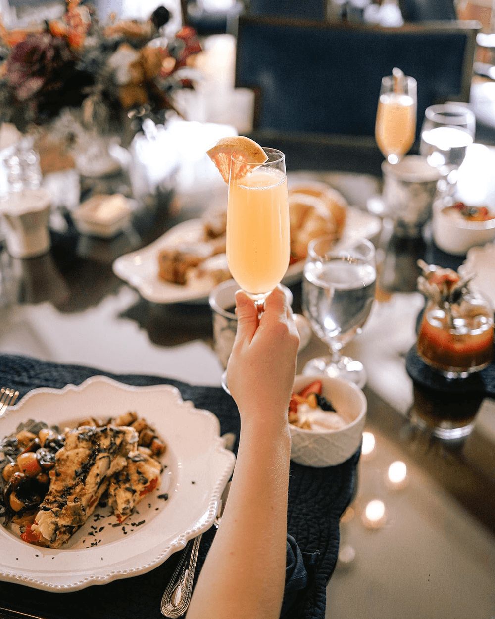 A hand raises a glass of mimosa over an elegant brunch spread on a reflective table.