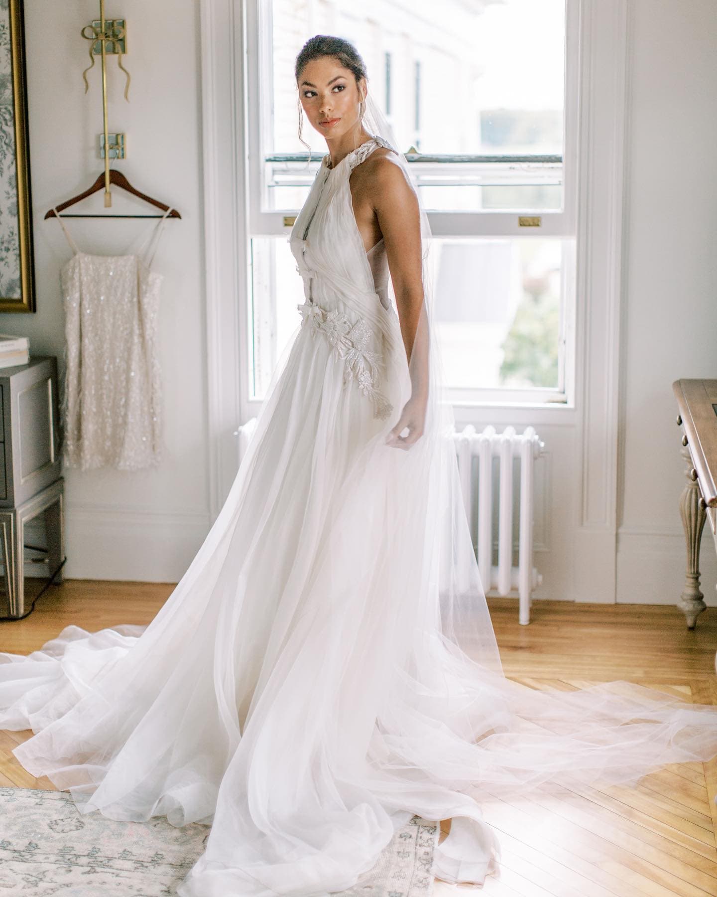 A woman in a flowing white gown stands in a bright room by a window, showcasing the dress's elegant design.