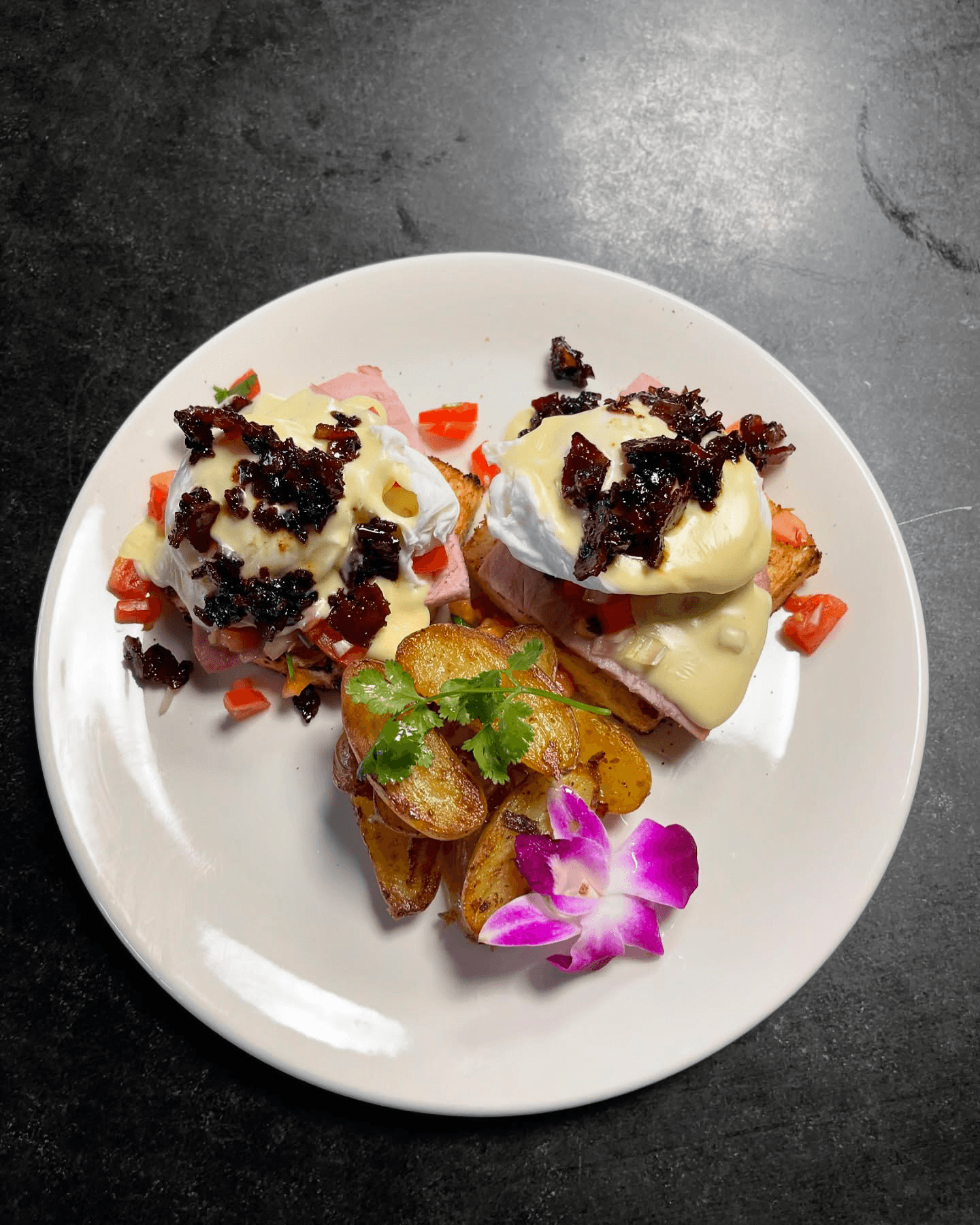 Two eggs Benedict with poached eggs, ham, hollandaise sauce, crispy toppings, roasted potatoes, and a purple flower garnish.