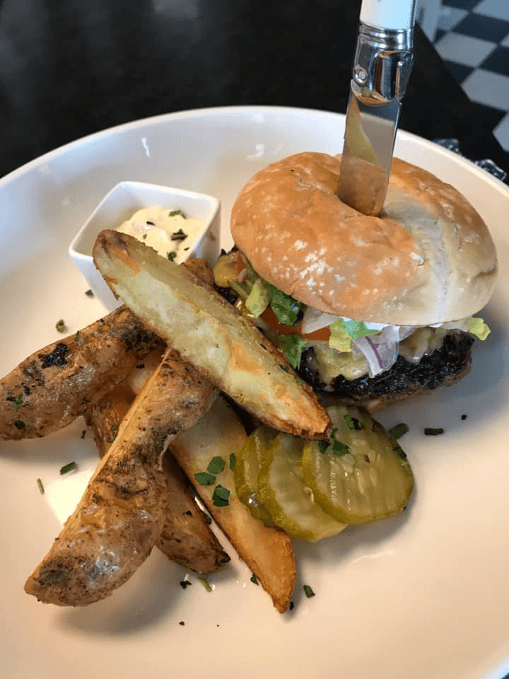 A burger with toppings and a knife inserted, accompanied by seasoned potato wedges and a small dish of dipping sauce.