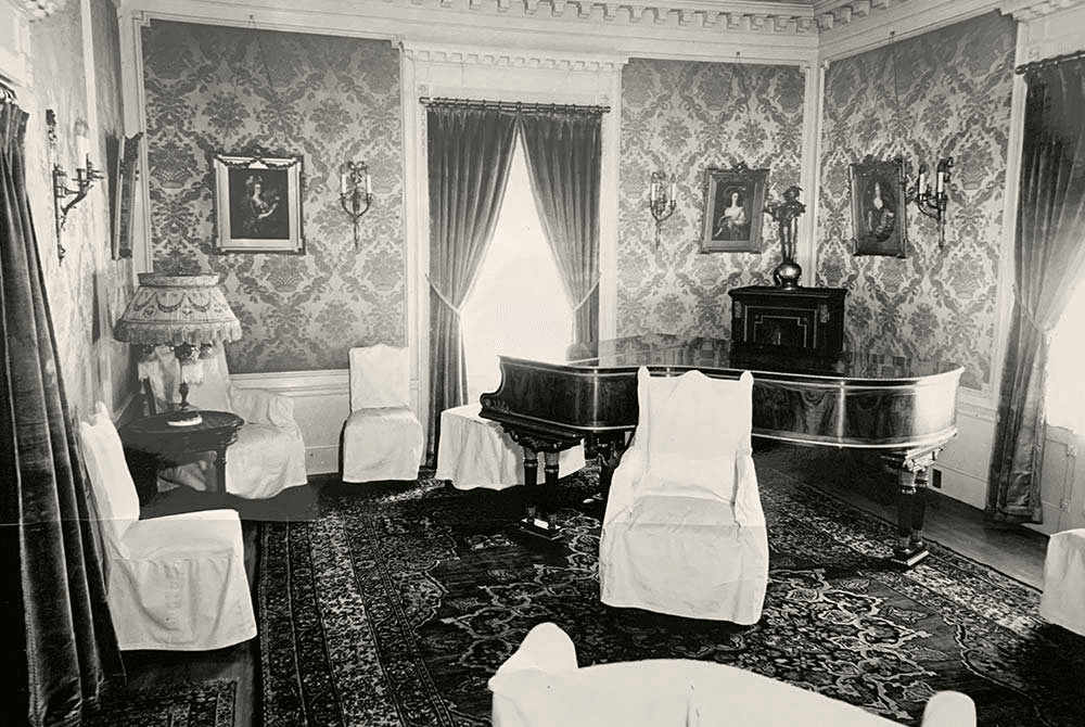 A vintage black and white photo of an elegant sitting room featuring a grand piano, draped windows, and upholstered chairs.
