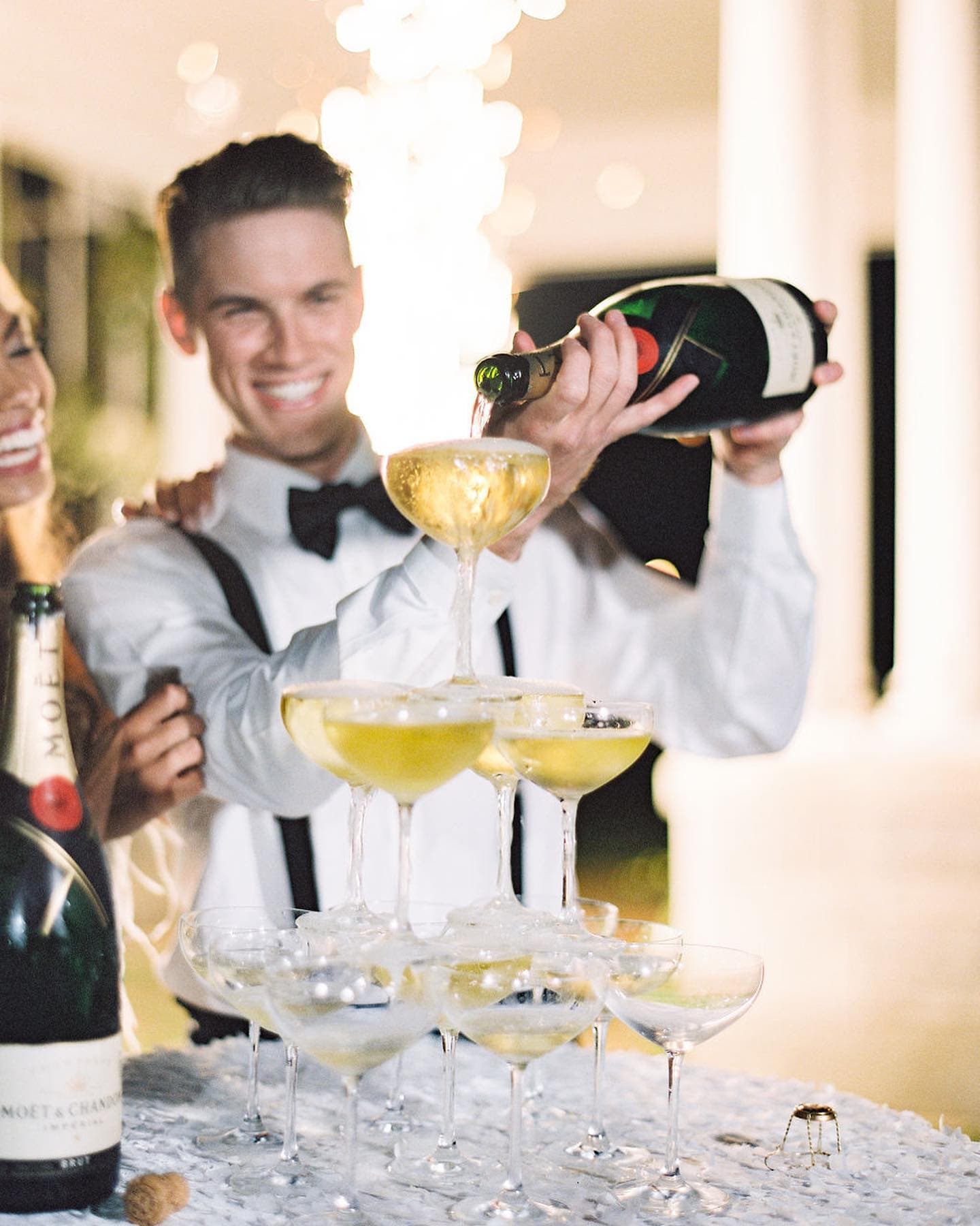 A smiling man in formal attire pours champagne into a pyramid of glasses at a festive event.