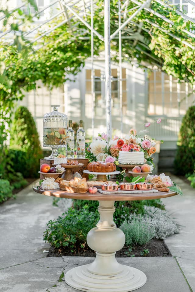 A beautifully arranged outdoor table featuring a variety of pastries, fruits, and floral decor under a vine-covered canopy.