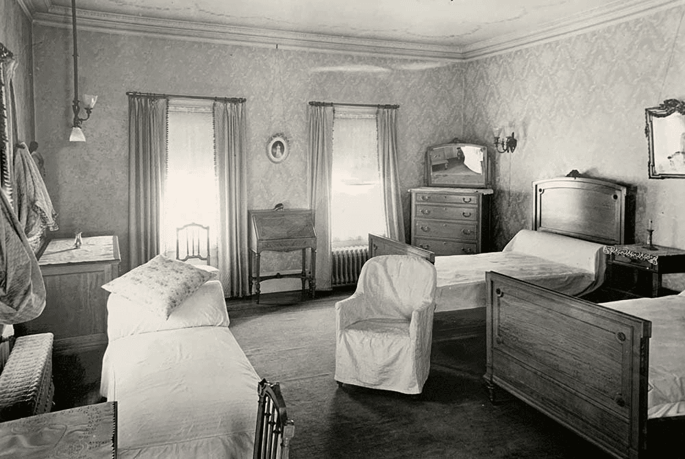 A vintage black-and-white photo of a bedroom with two beds, a chair, and a dresser, featuring patterned wallpaper and window curtains.
