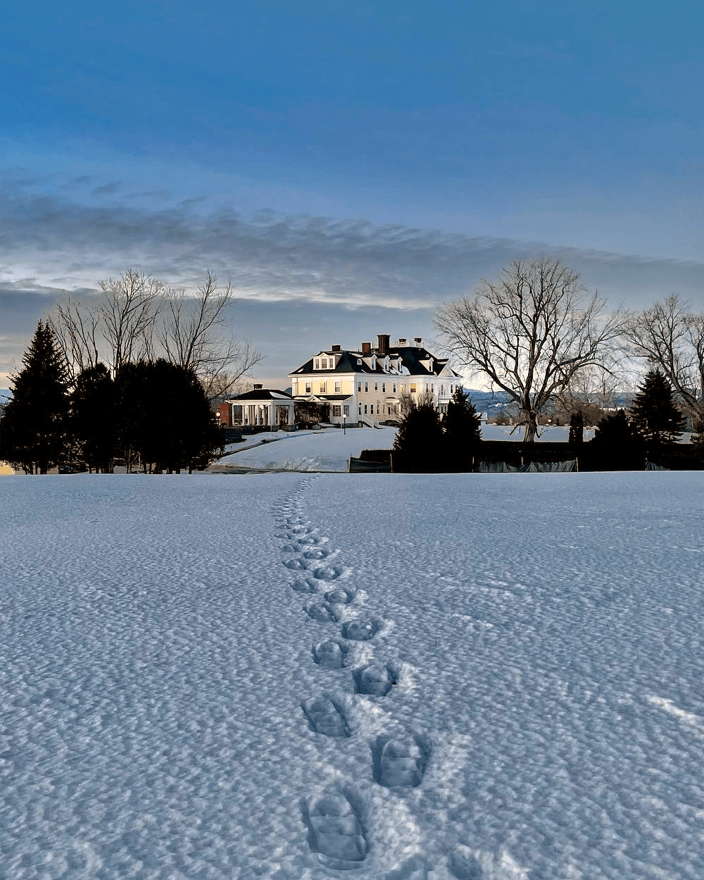 Footprints lead through snow towards a large, illuminated mansion at dusk.