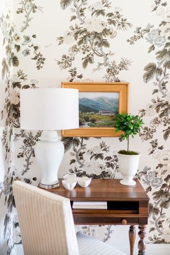 A wooden table with a white lamp, small plant, and dishware against a floral-patterned wall.