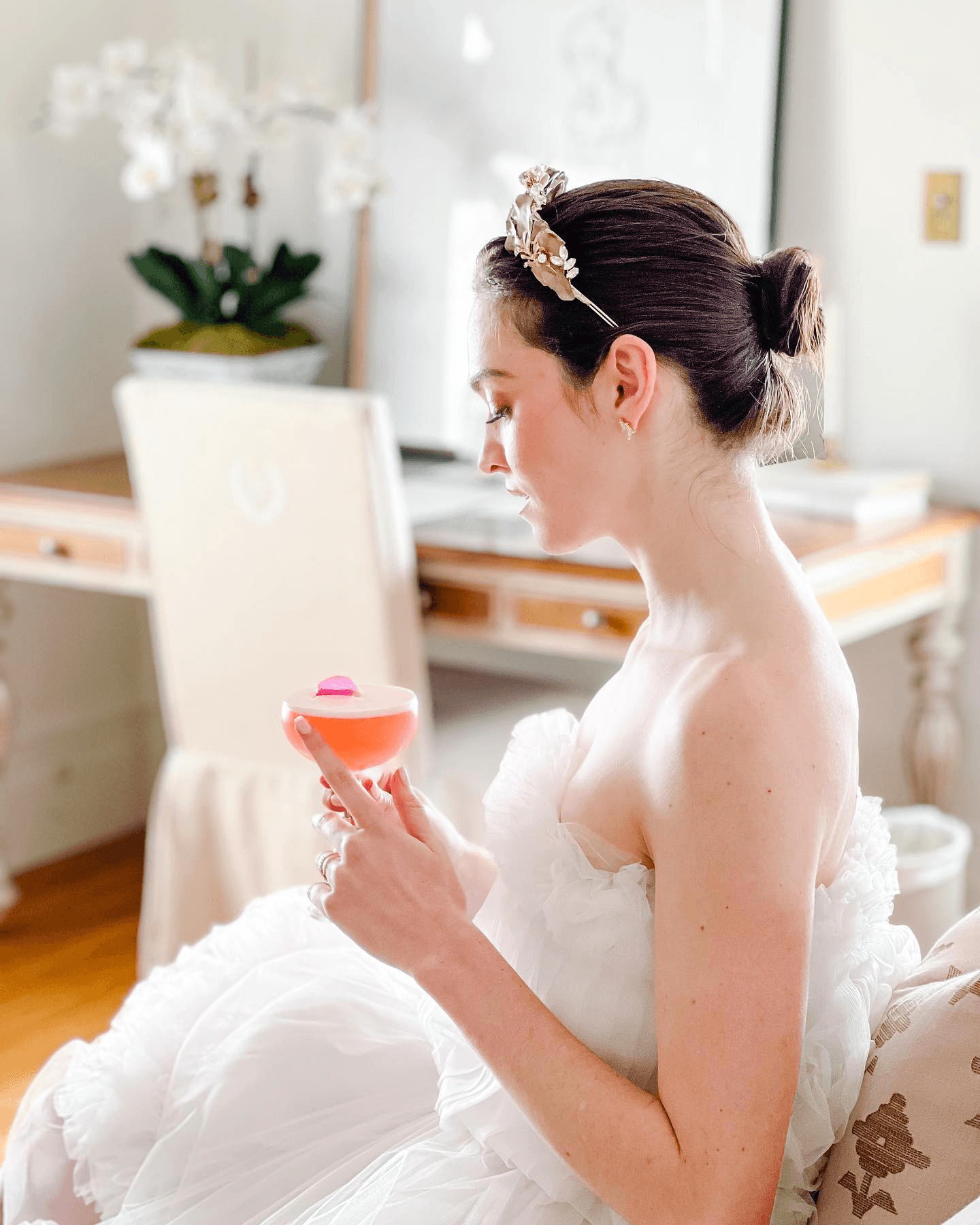 A woman in a strapless white dress holds a cocktail while sitting in a well-decorated room.