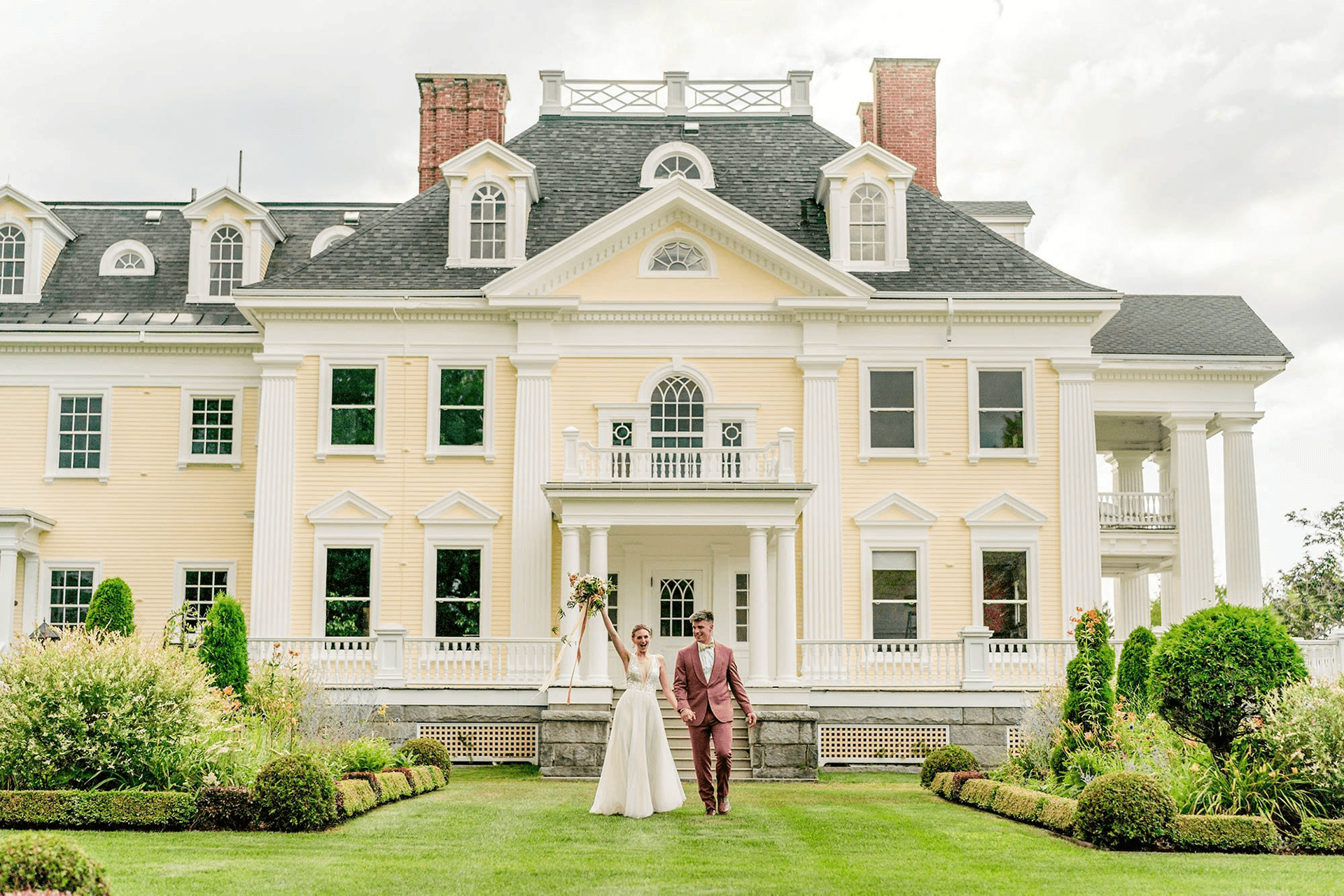 A newlywed couple walks hand in hand in front of a large yellow mansion surrounded by greenery.