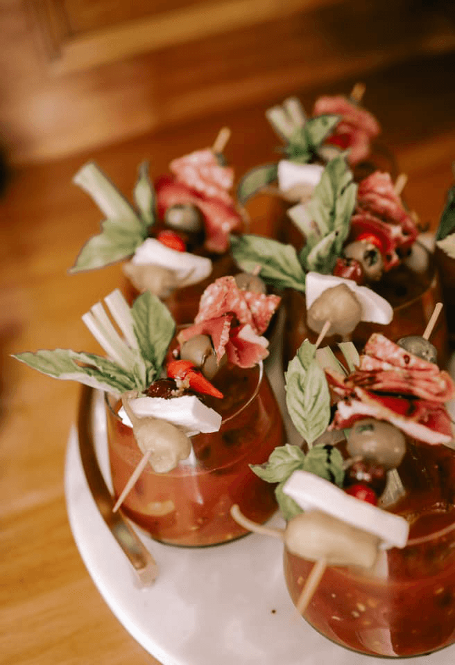A close-up of cocktails garnished with meats, olives, and fresh herbs on a wooden surface.