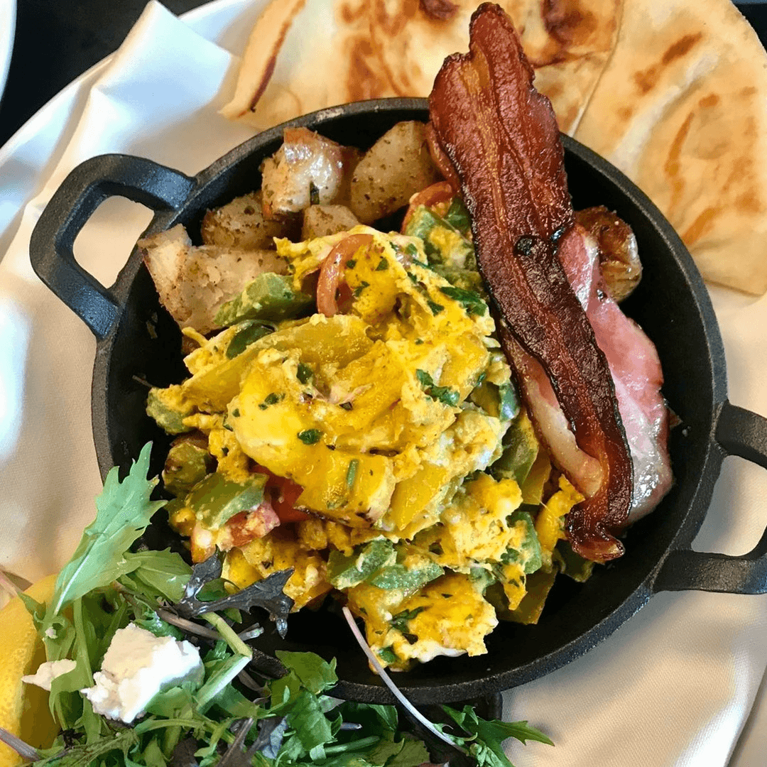 A cast iron skillet filled with scrambled eggs, mixed vegetables, bacon, roasted potatoes, and a side of greens.