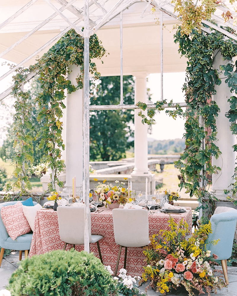 A beautifully decorated dining table with vibrant floral arrangements and greenery set within a glass-enclosed space.