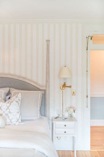 A stylish bedroom featuring a gray tufted headboard, patterned pillows, a bedside table with a lamp and flowers, and soft striped walls.