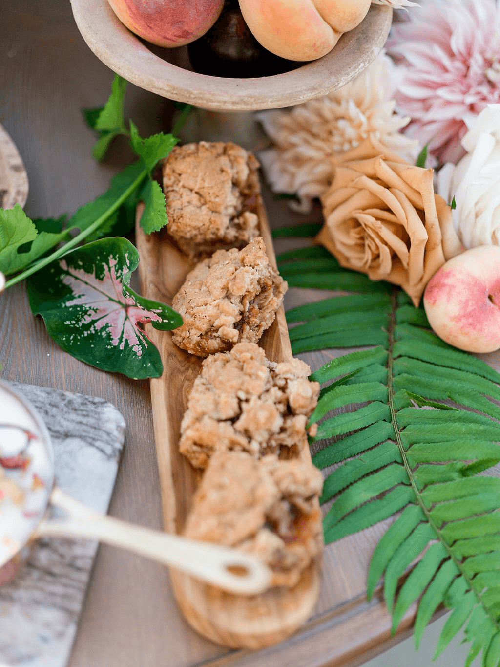 A wooden platter holds crumbly baked goodies, surrounded by fresh peaches, roses, and green foliage.