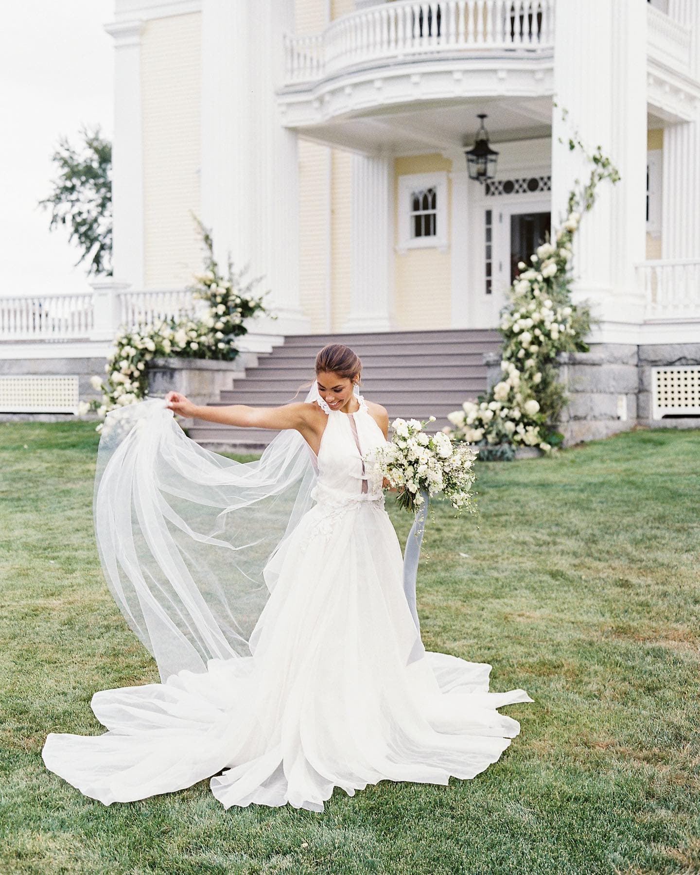 A smiling bride in a flowing white gown twirls with her veil and bouquet in front of a beautifully decorated venue.