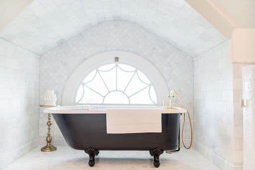 A black clawfoot bathtub sits elegantly under a large, arched window in a marble-tiled bathroom.