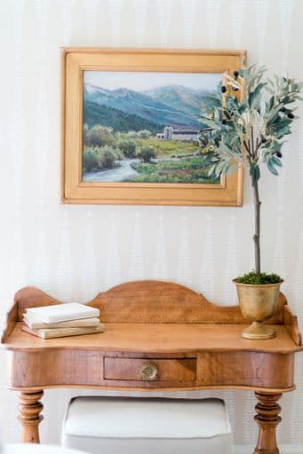 A wooden desk with books and a small potted plant sits below a framed landscape painting on a textured wall.
