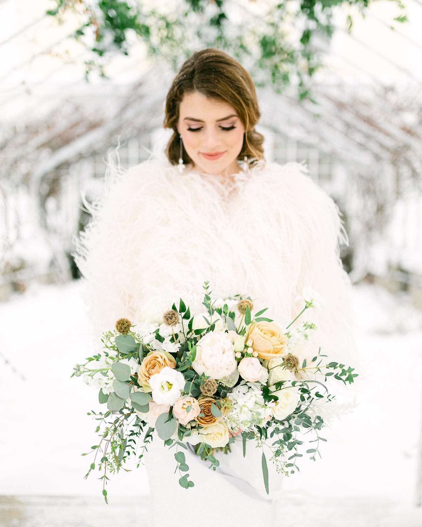 A woman in a fluffy white shawl holds a bouquet of soft-colored flowers in a snowy setting.