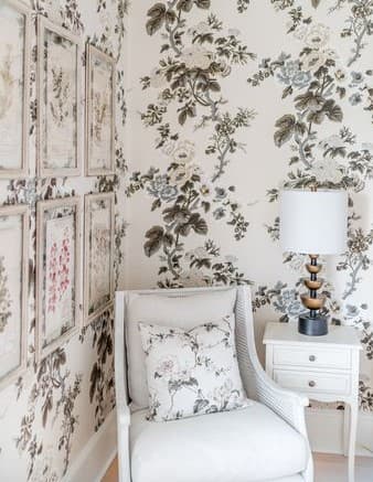A cozy corner featuring a white chair with floral cushions, a small table, and a lamp against a patterned floral wallpaper.