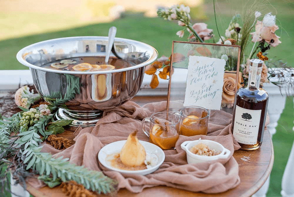 A silver punch bowl with fruit slices, two glasses of punch, a dessert, a bottle of whiskey, and a decorative sign are arranged on a wooden table adorned with flowers and greenery.