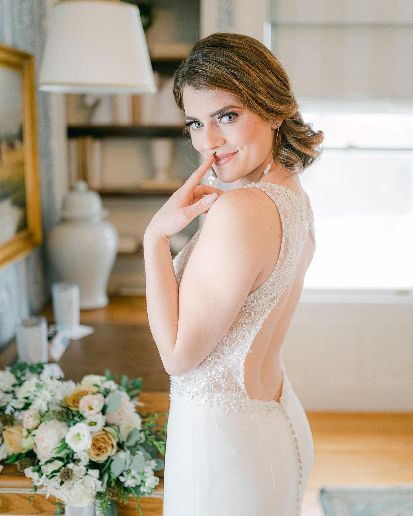 A bride in a backless wedding gown playfully gestures with her finger while posing in a beautifully decorated room.