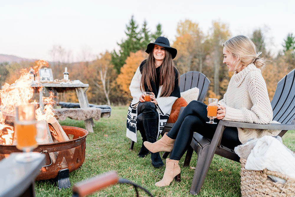 Two women enjoy drinks by a campfire surrounded by autumn foliage.