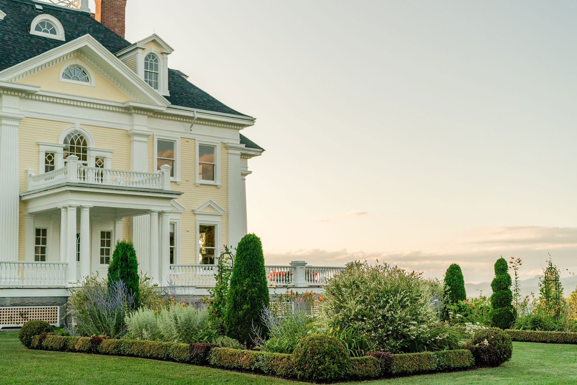 A grand yellow house with an ornate facade surrounded by neatly trimmed gardens under a clear sky.