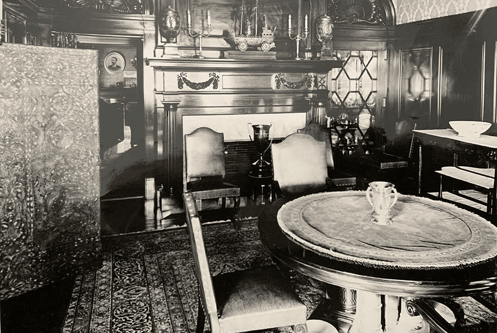A vintage black-and-white photo of an elegantly furnished room featuring a round table, upholstered chairs, and ornate decor.