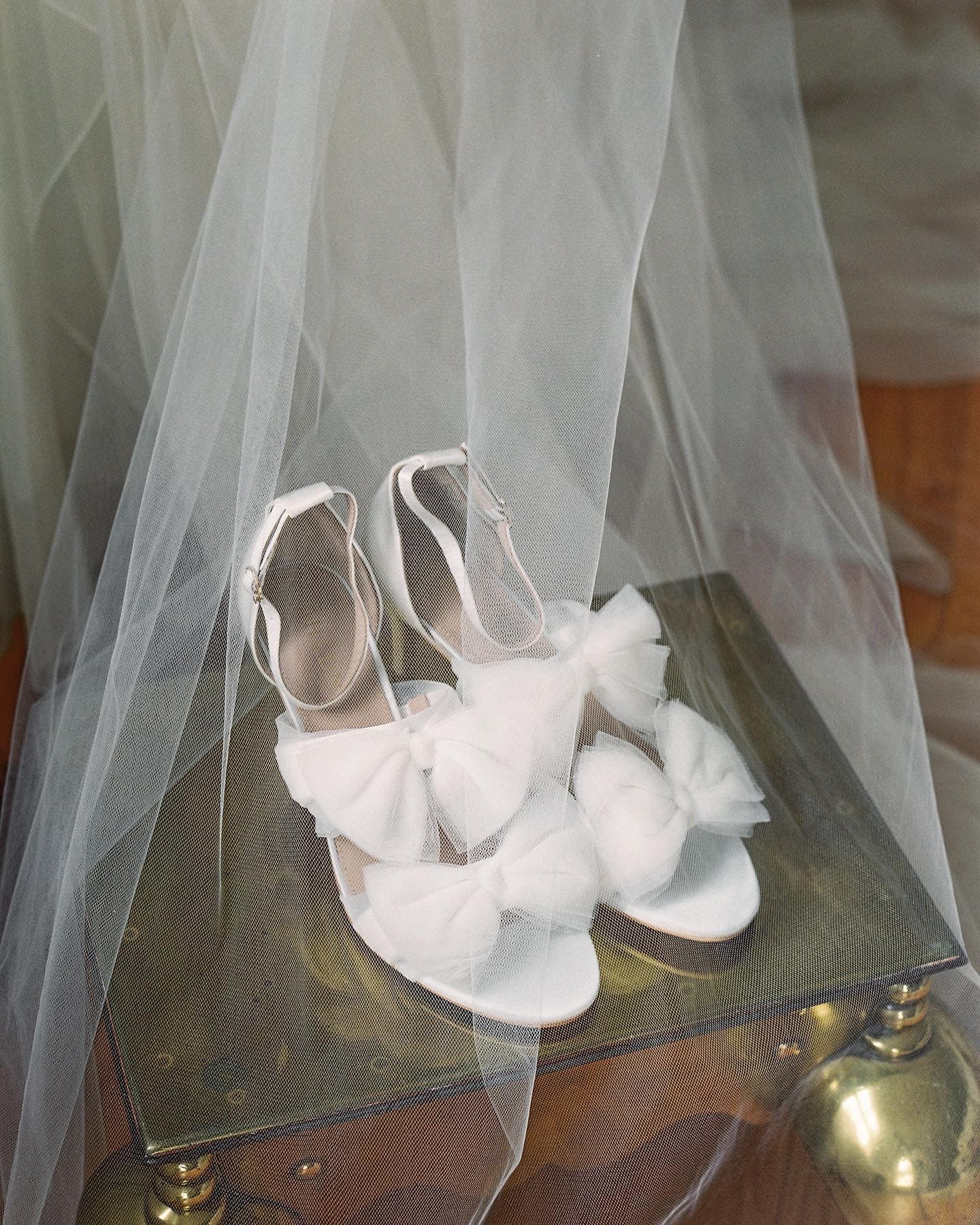 White shoes with tulle bows resting on a table covered by a sheer veil.